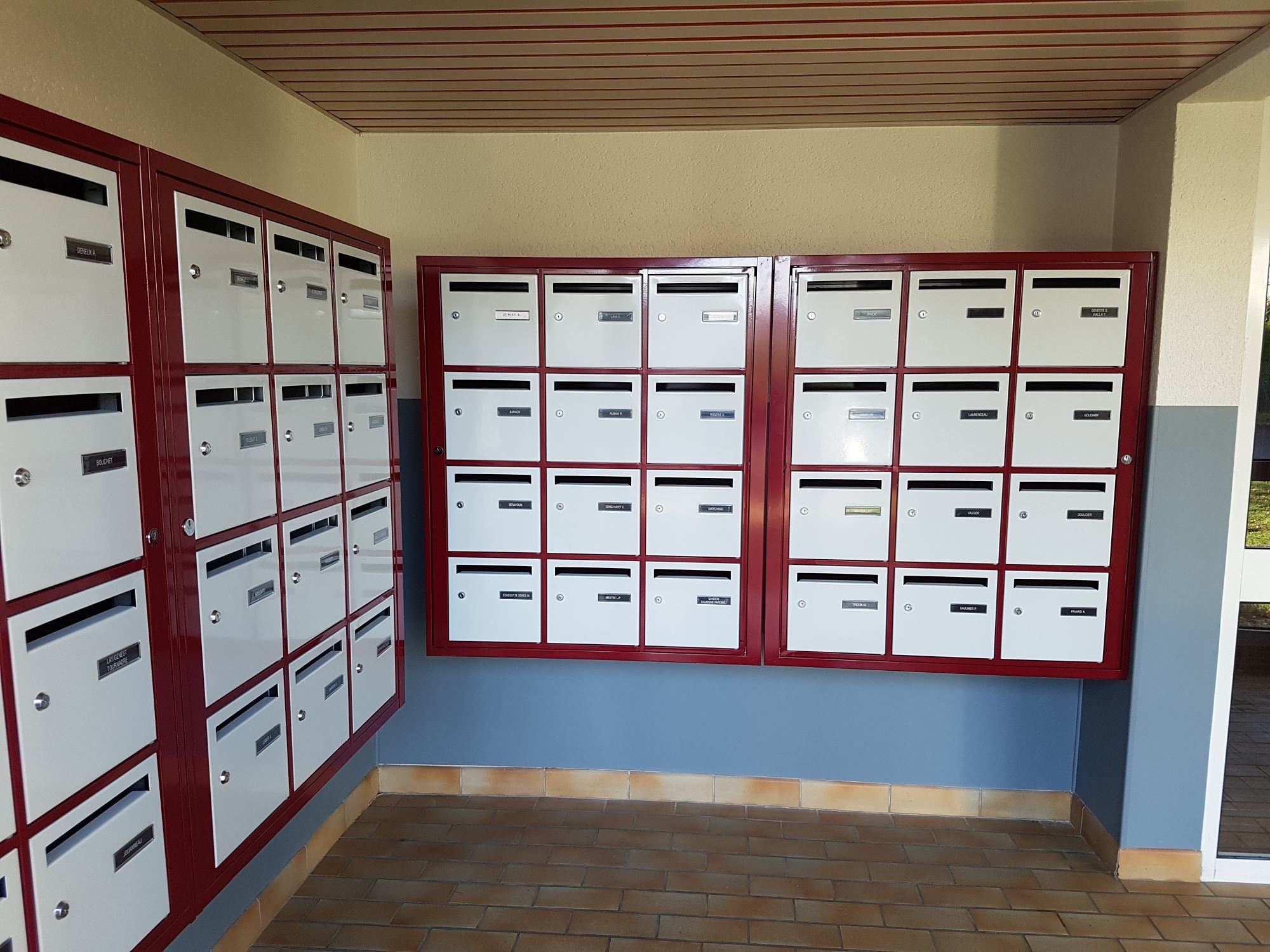 Des ensembles de boîtes aux lettres blanches encadrées de rouge sont fixés aux murs d'un hall d'entrée couvert au sol carrelé.