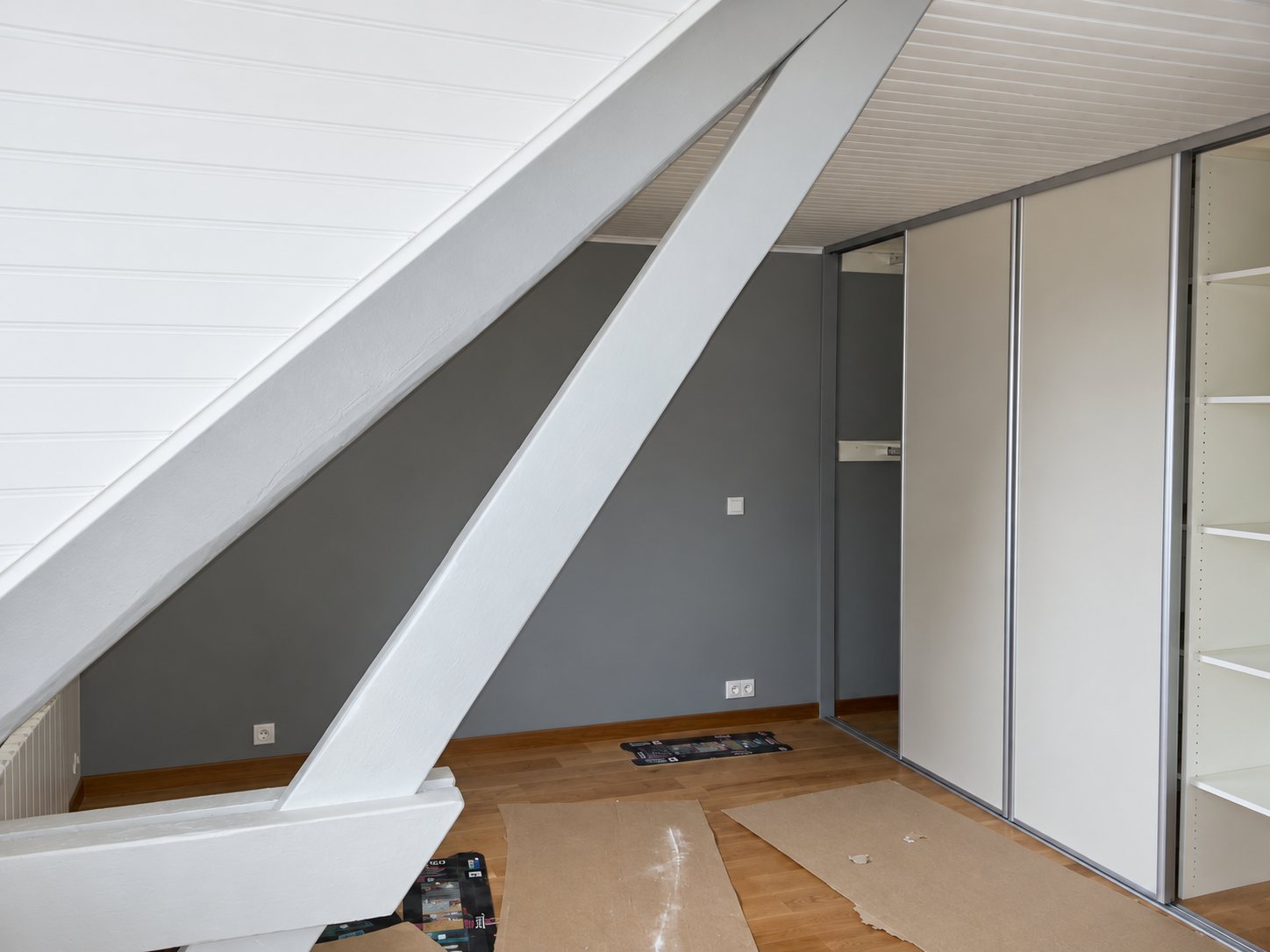Chambre mansardée aux murs gris et poutres blanches, équipée d'un placard à portes coulissantes, avec des cartons de protection posés sur le parquet.