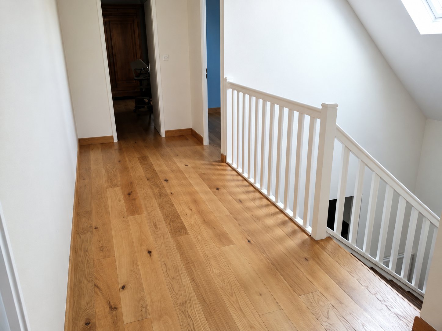 Un couloir à l'étage au parquet de chêne clair fraîchement posé, bordé d'une rampe d'escalier blanche et de murs blancs.