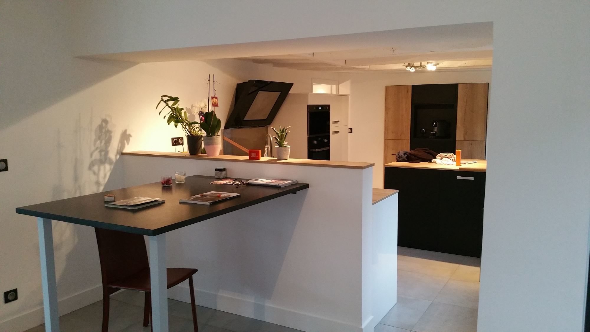 Cuisine contemporaine ouverte avec façades noires et bois clair, hotte inclinée, bar blanc surmonté de plantes et table haute noire avec magazines.