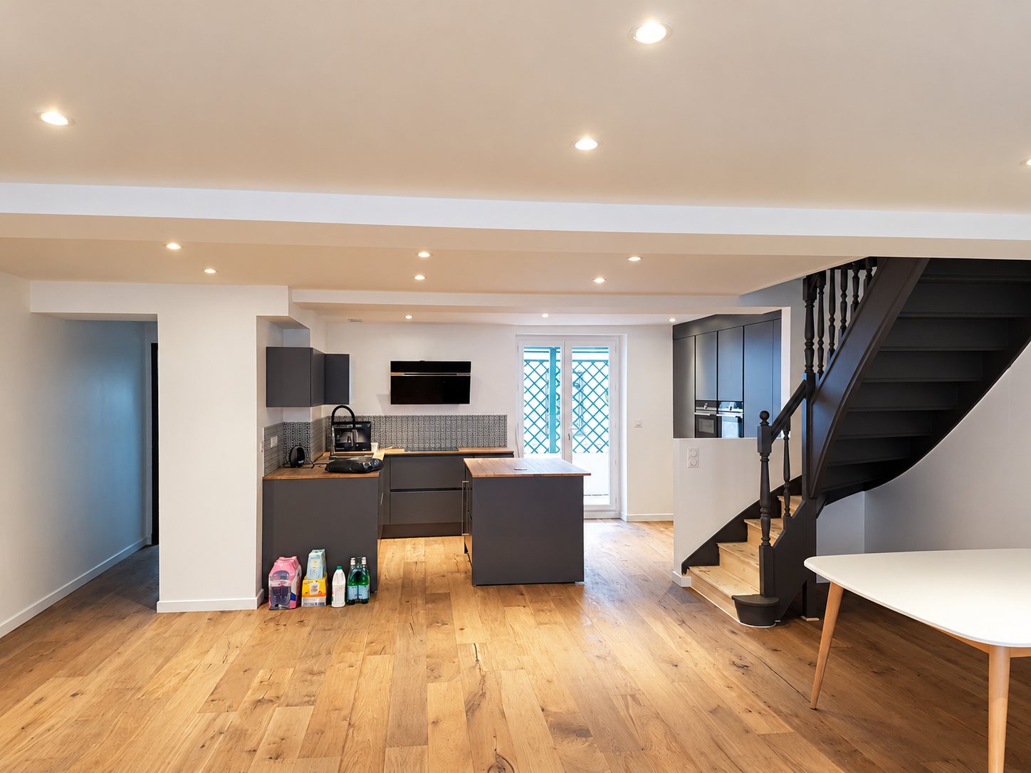 Pièce de vie rénovée avec cuisine grise ouverte et îlot central, parquet en chêne clair, spots encastrés et escalier en bois peint en gris foncé.