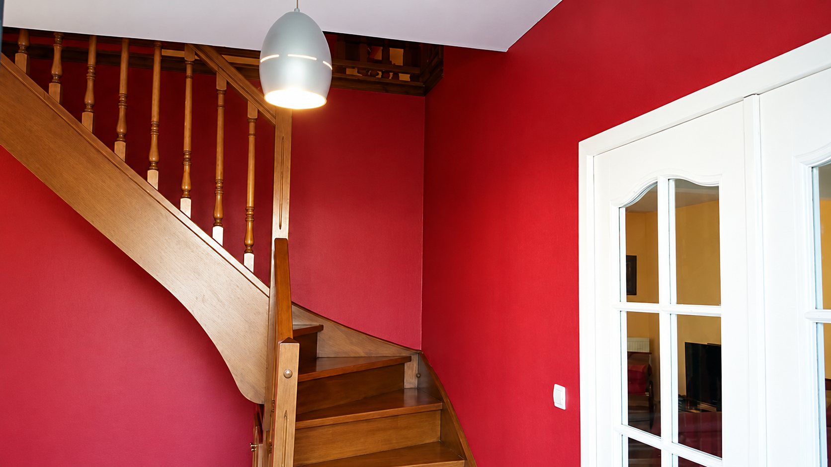 Un escalier tournant en bois avec balustres, entouré de murs rouges, une suspension grise et une porte blanche vitrée.