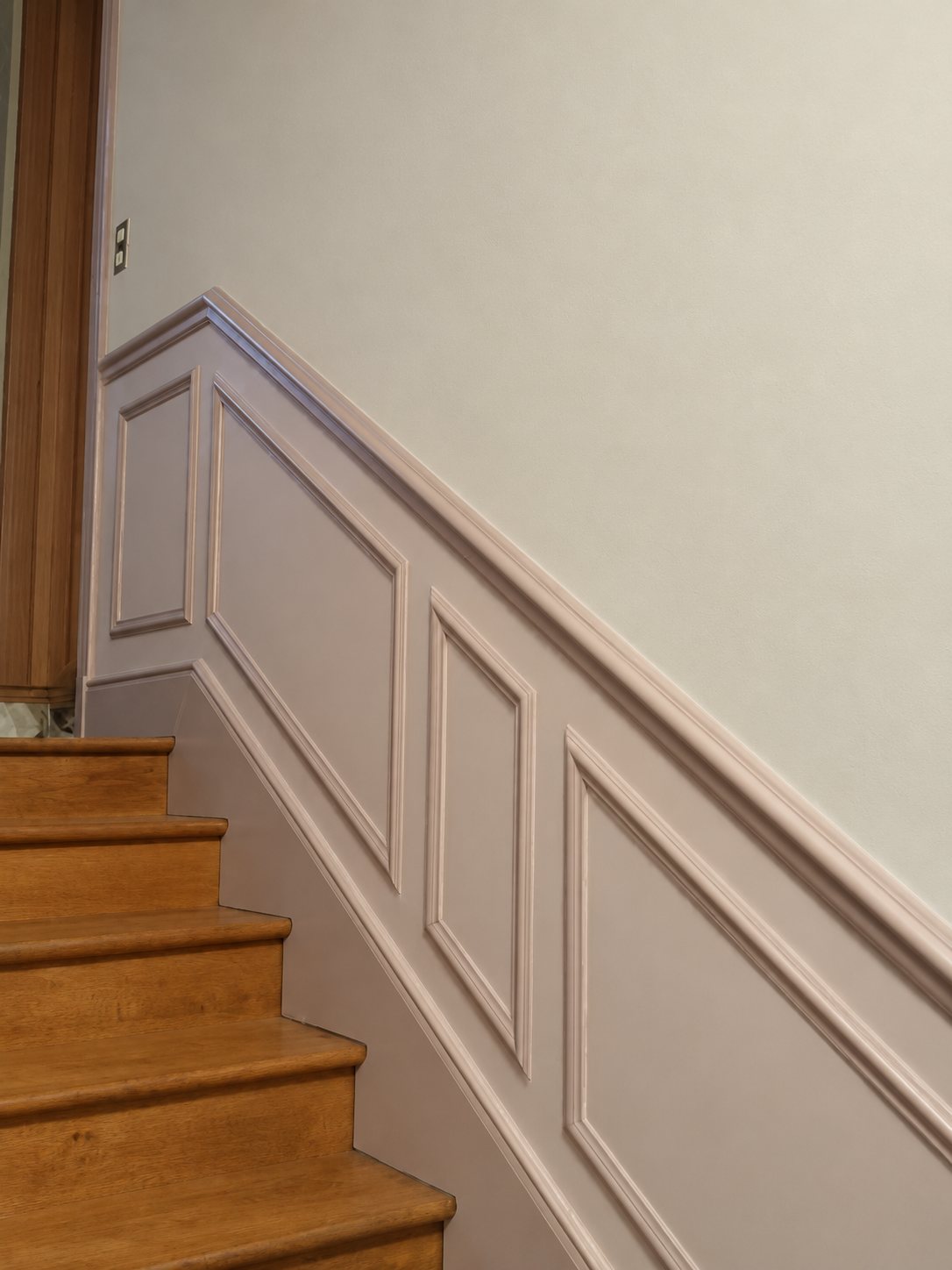 Un escalier aux marches en bois longé par un soubassement à panneaux moulurés peint en rose pâle, sous un mur clair.