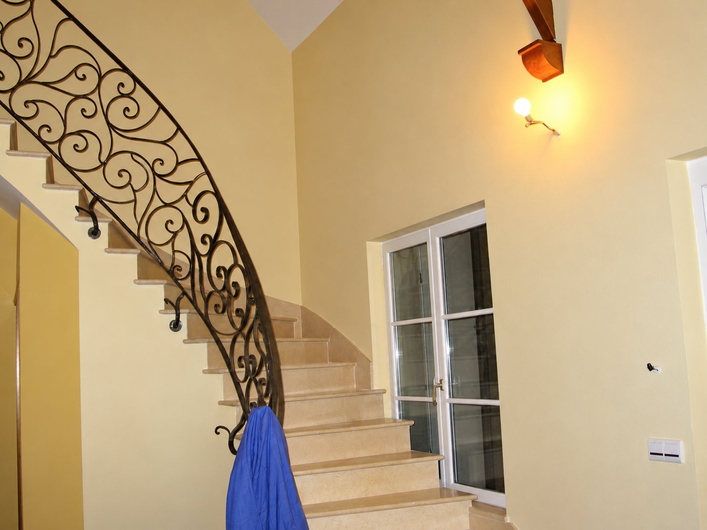 Un escalier courbe en pierre claire avec une rampe en fer forgé à volutes, dans une entrée aux murs jaune pâle près d'une porte-fenêtre blanche.