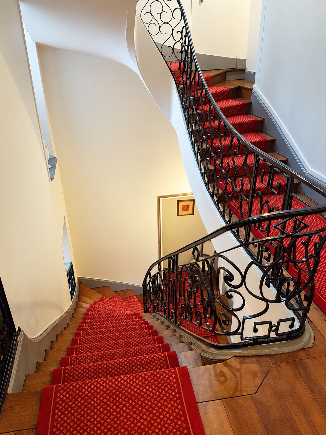 Vue plongeante sur un escalier tournant aux murs blancs, doté d'une rampe en fer forgé noir et d'un tapis rouge à motifs.
