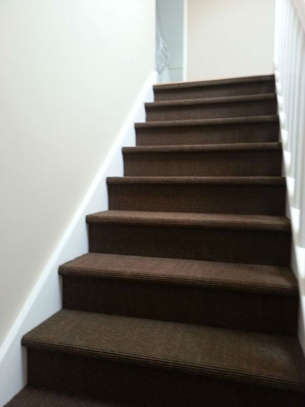Un escalier droit dont les marches sont recouvertes d'une moquette côtelée marron foncé, bordé d'un mur beige et d'une rampe blanche.