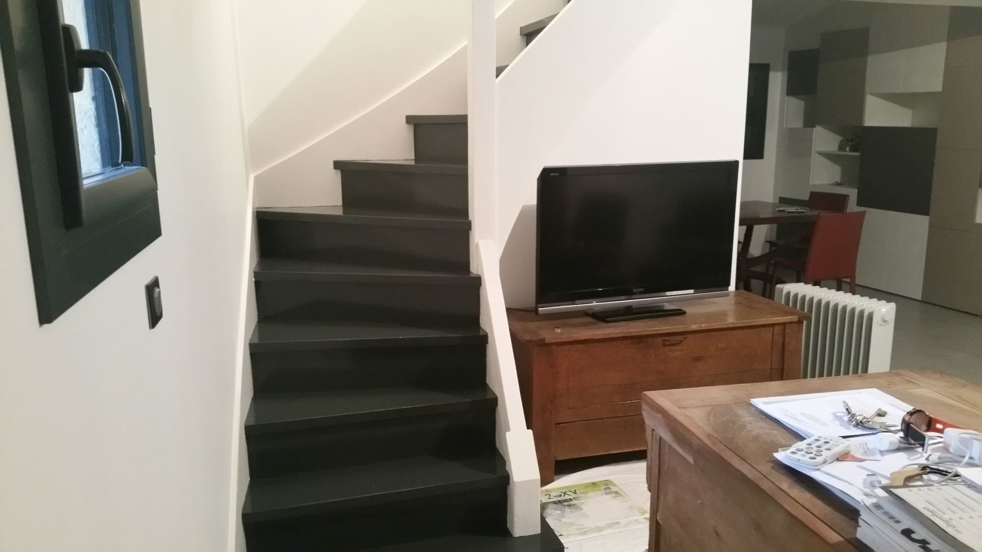 Un escalier tournant aux marches gris anthracite et aux murs blancs, à côté d'un meuble en bois avec un téléviseur, dans un séjour rénové.