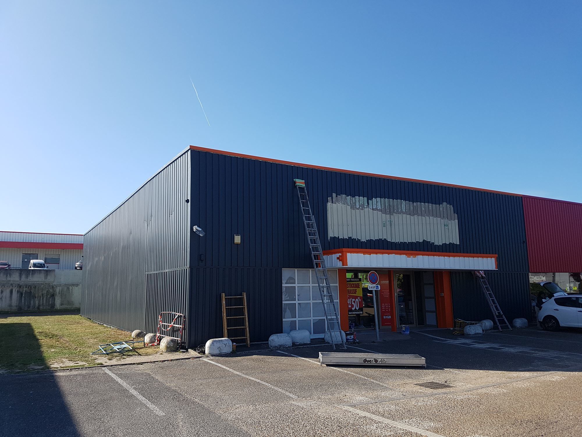 Façade d'un magasin en bardage métallique gris foncé avec liserés orange, où deux échelles sont appuyées au mur et où l'ancienne enseigne a été déposée.