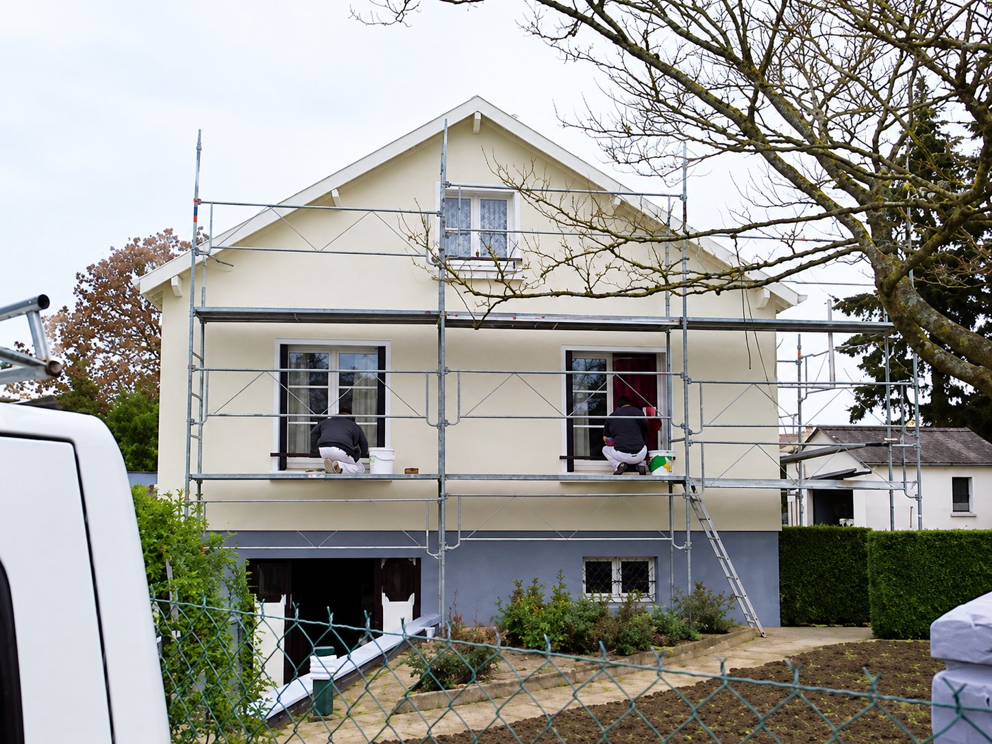 Une maison à façade crème et soubassement bleu-gris entourée d'un échafaudage, où deux peintres travaillent aux fenêtres.