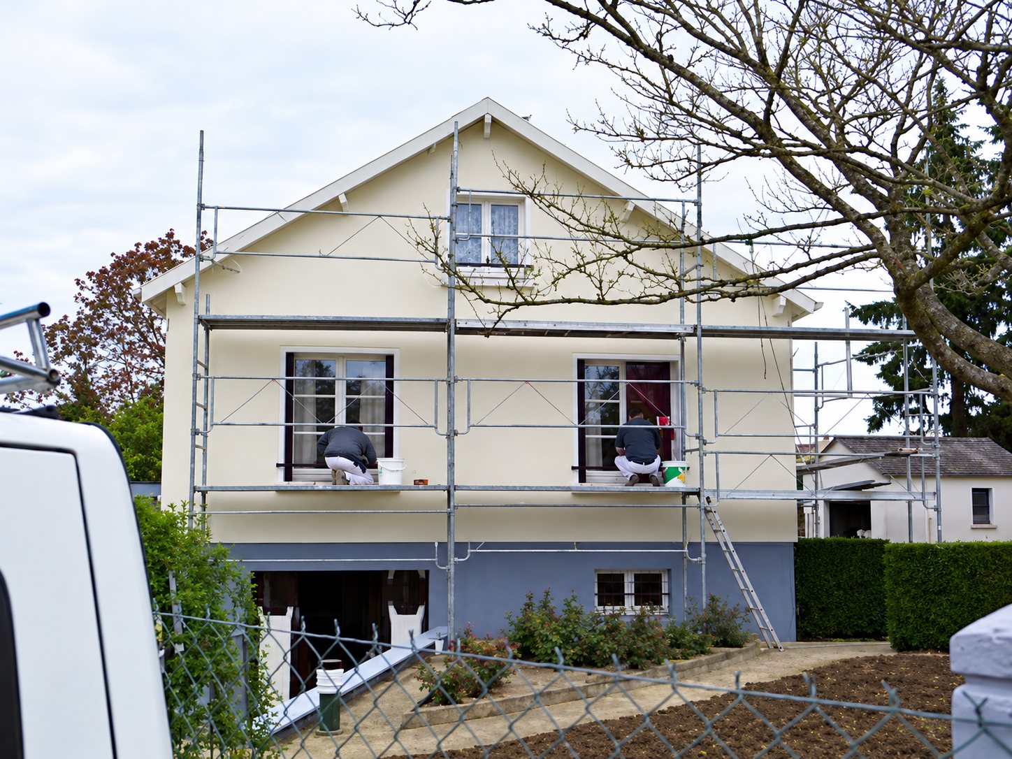 Une maison à pignon à la façade crème est entourée d'un échafaudage, tandis que deux ouvriers travaillent aux fenêtres de l'étage.