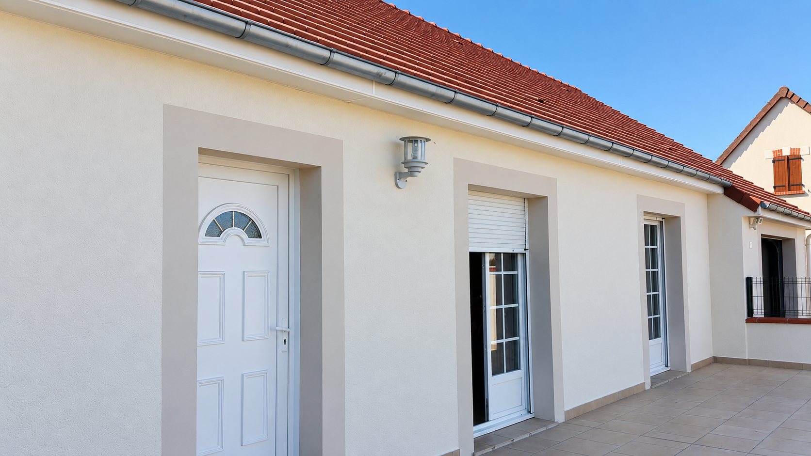 Façade d'une maison de plain-pied fraîchement enduite en crème, avec encadrements beiges autour de la porte d'entrée blanche et des fenêtres, toit en tuiles rouges et terrasse carrelée.
