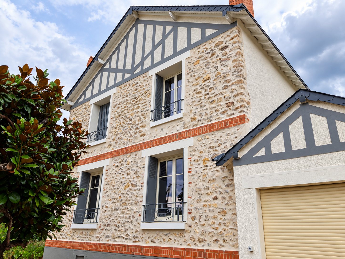 Façade rénovée d'une maison en pierre meulière avec faux colombages gris, bandeaux de briques orange, volets gris et garage à porte beige.