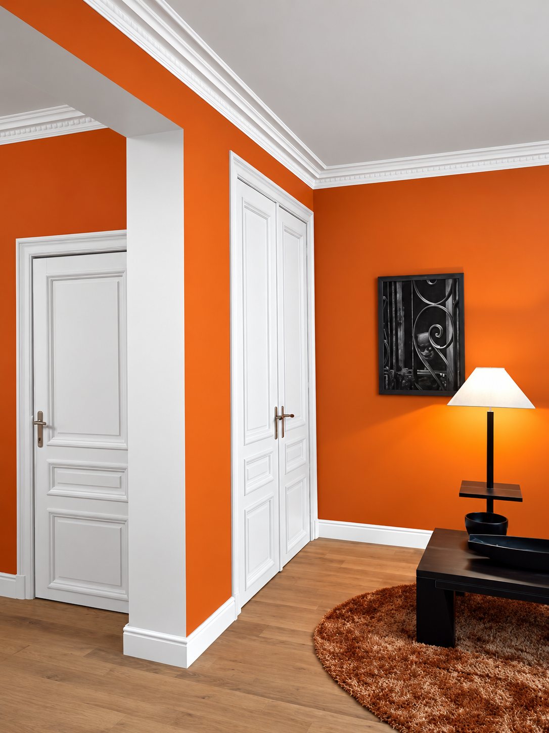 Un salon aux murs orange vif avec des portes blanches à panneaux, des corniches blanches au plafond, un parquet clair, un tapis rond brun et une lampe sur une table basse sombre.