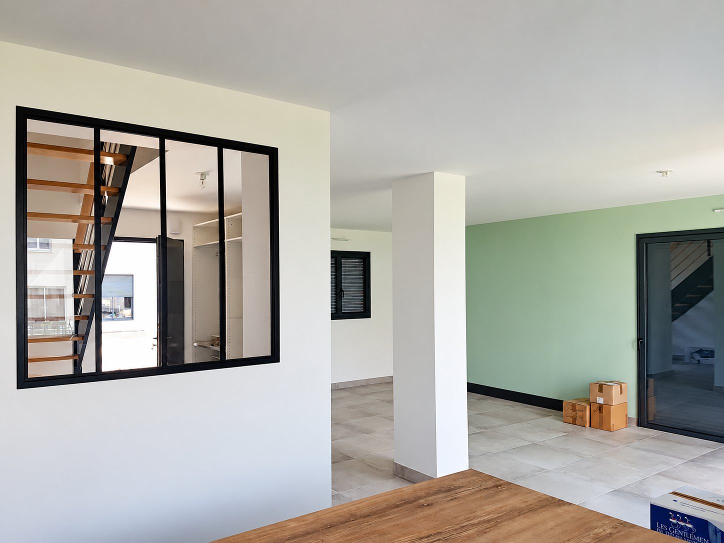 Pièce de vie spacieuse récemment rénovée avec une verrière d'atelier noire, un pilier blanc, un mur vert d'eau, un carrelage gris et quelques cartons au sol.
