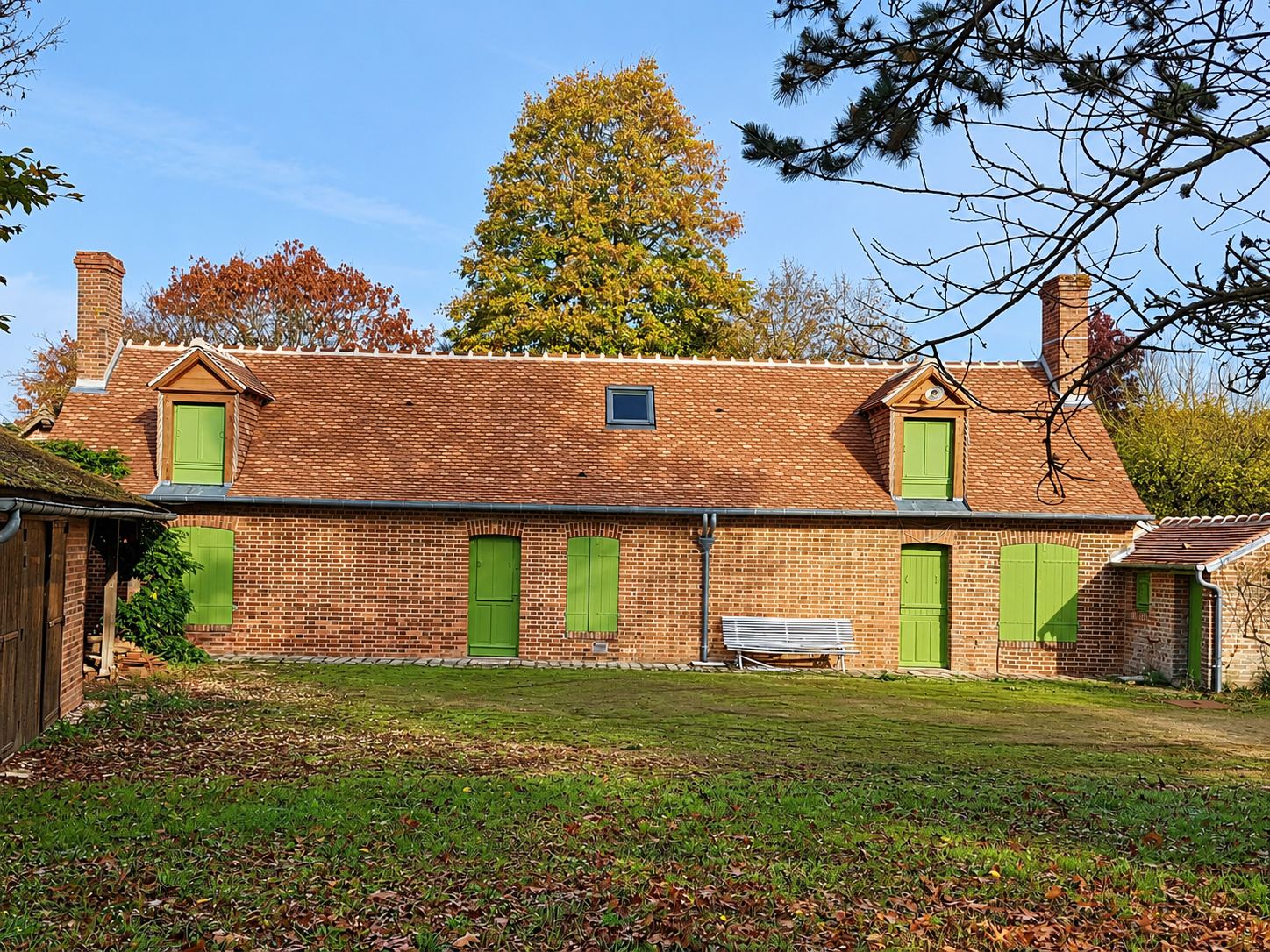 Longue maison en brique rouge aux volets et portes verts, avec un toit de tuiles flammées neuf et des lucarnes en bois, entourée d'arbres d'automne.