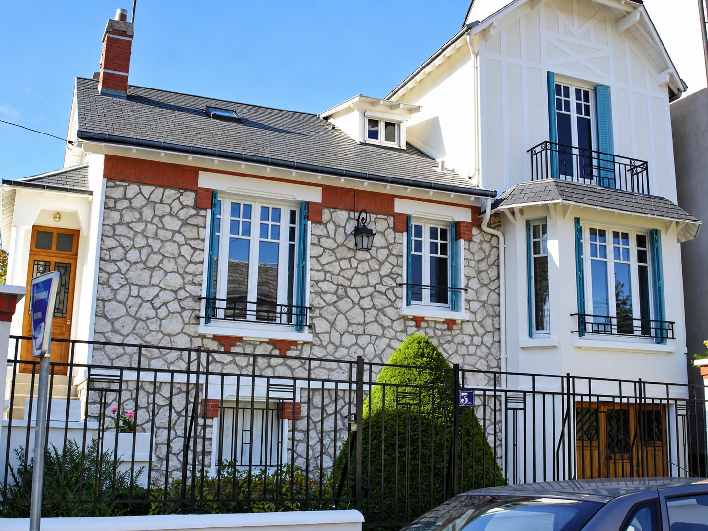 Façade d'une maison ancienne en pierre et crépi blanc, avec volets bleus, balcon en fer forgé, portail noir et un petit buis taillé devant.