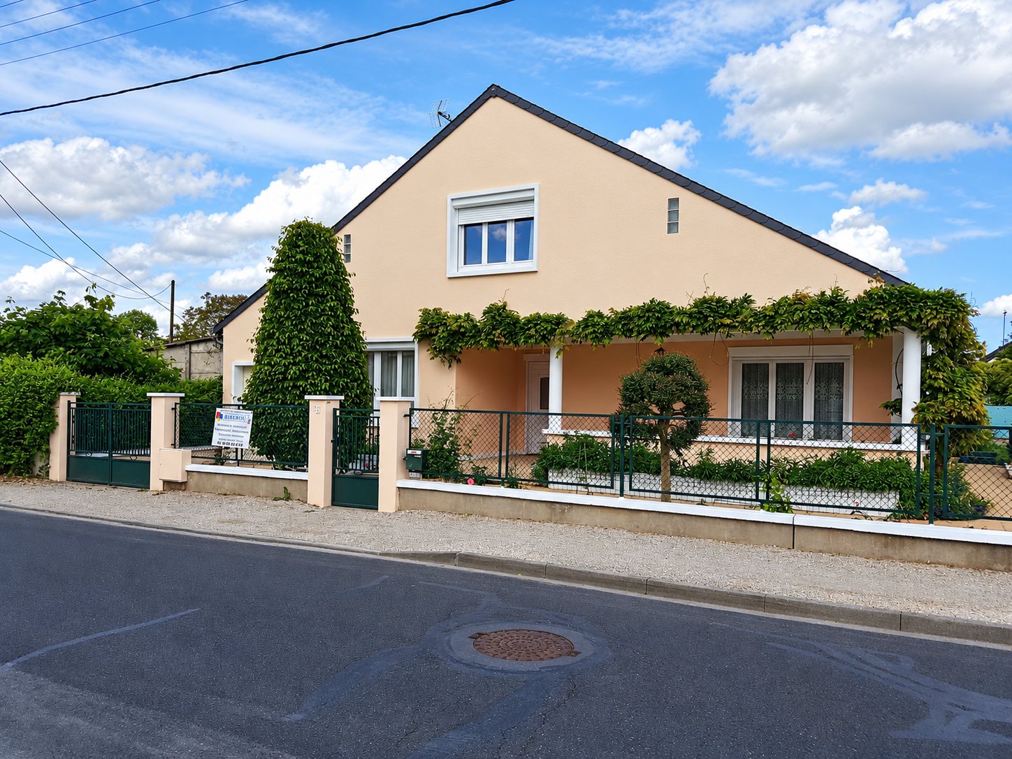 Maison individuelle à façade rose clair et toit sombre, avec véranda couverte de vigne, clôture, portail vert et petit panneau d'entreprise devant la rue.
