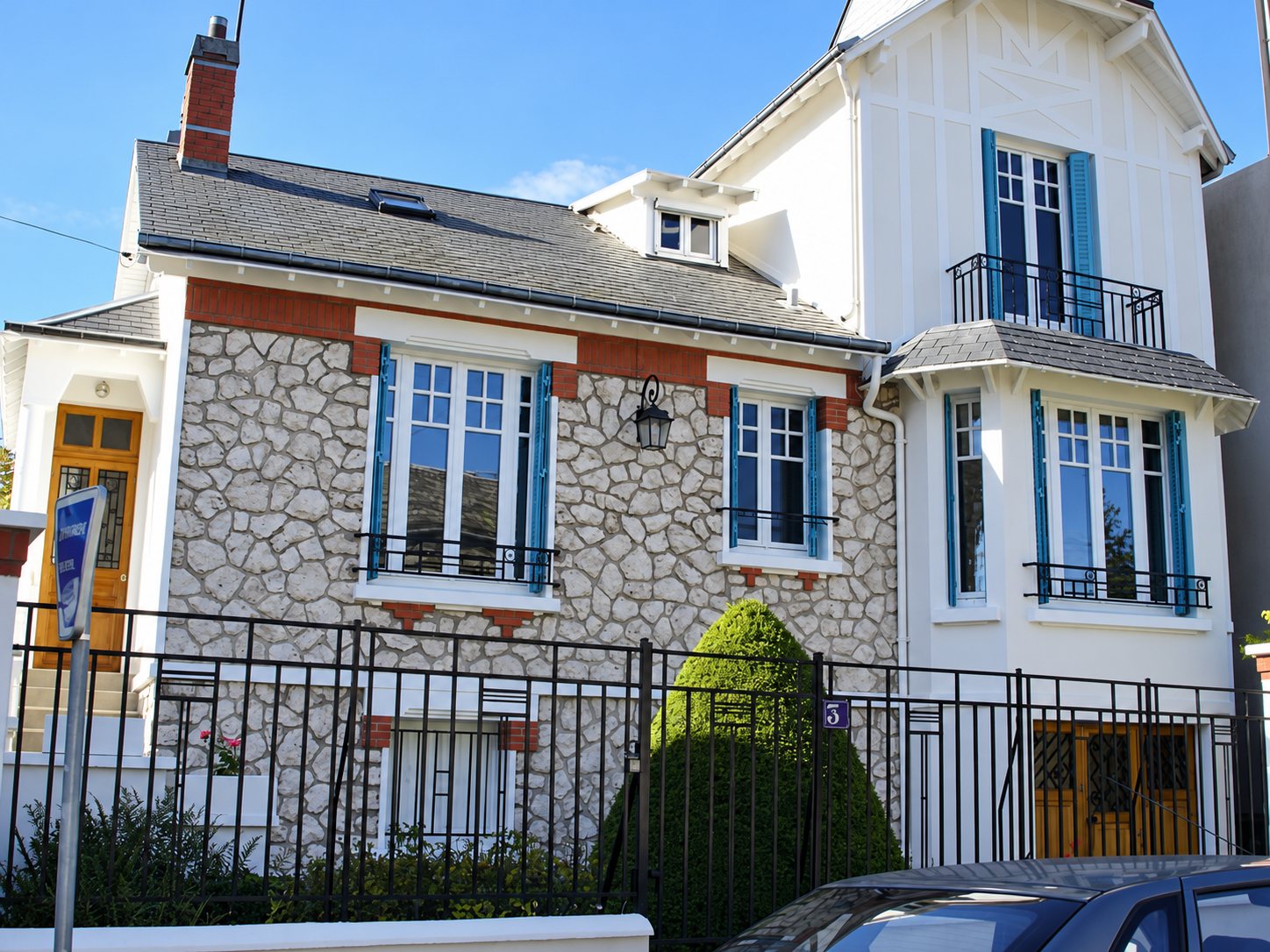 Façade d'une maison ancienne rénovée, avec murs en pierre apparente, encadrements en brique, volets bleus et balcon en fer forgé derrière un portail noir.