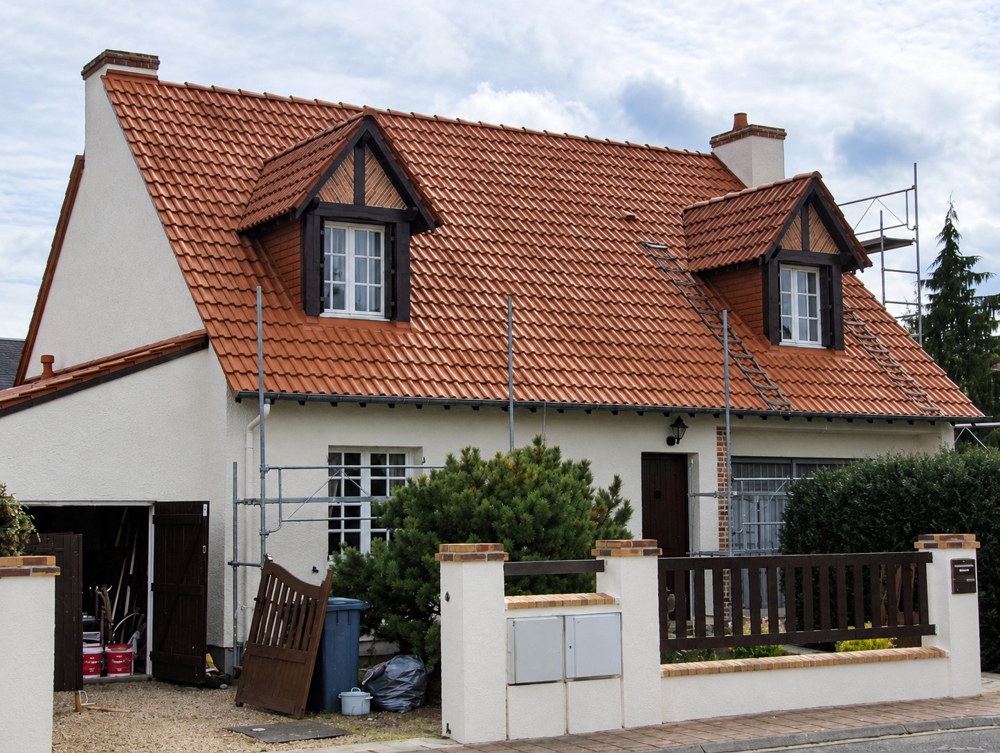 Maison blanche à deux lucarnes avec toit en tuiles rousses, entourée d'un échafaudage pendant des travaux de rénovation.