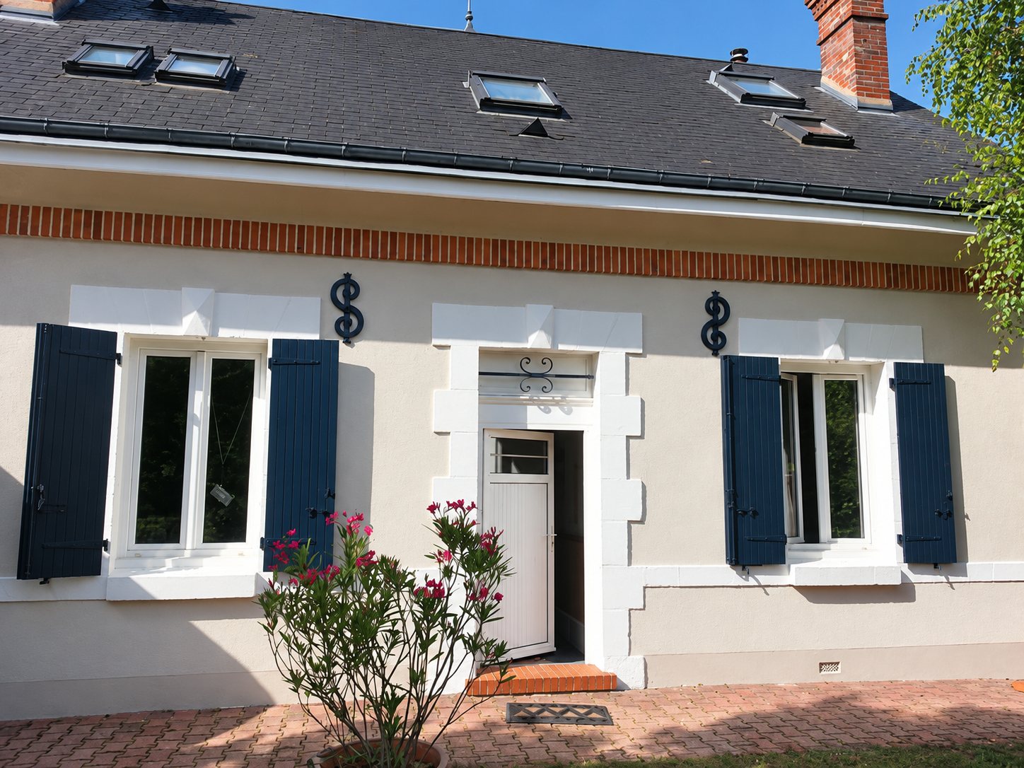 Façade d'une maison de plain-pied rénovée, avec enduit beige, volets bleu foncé, fenêtres et porte blanches, toit en ardoise avec fenêtres de toit et un laurier rose au premier plan.