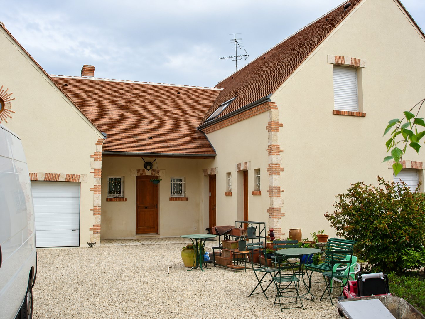 Façade d'une maison crépie beige avec toit en tuiles brunes, porte en bois et garage blanc, devant une cour de gravier meublée de tables et chaises de jardin.