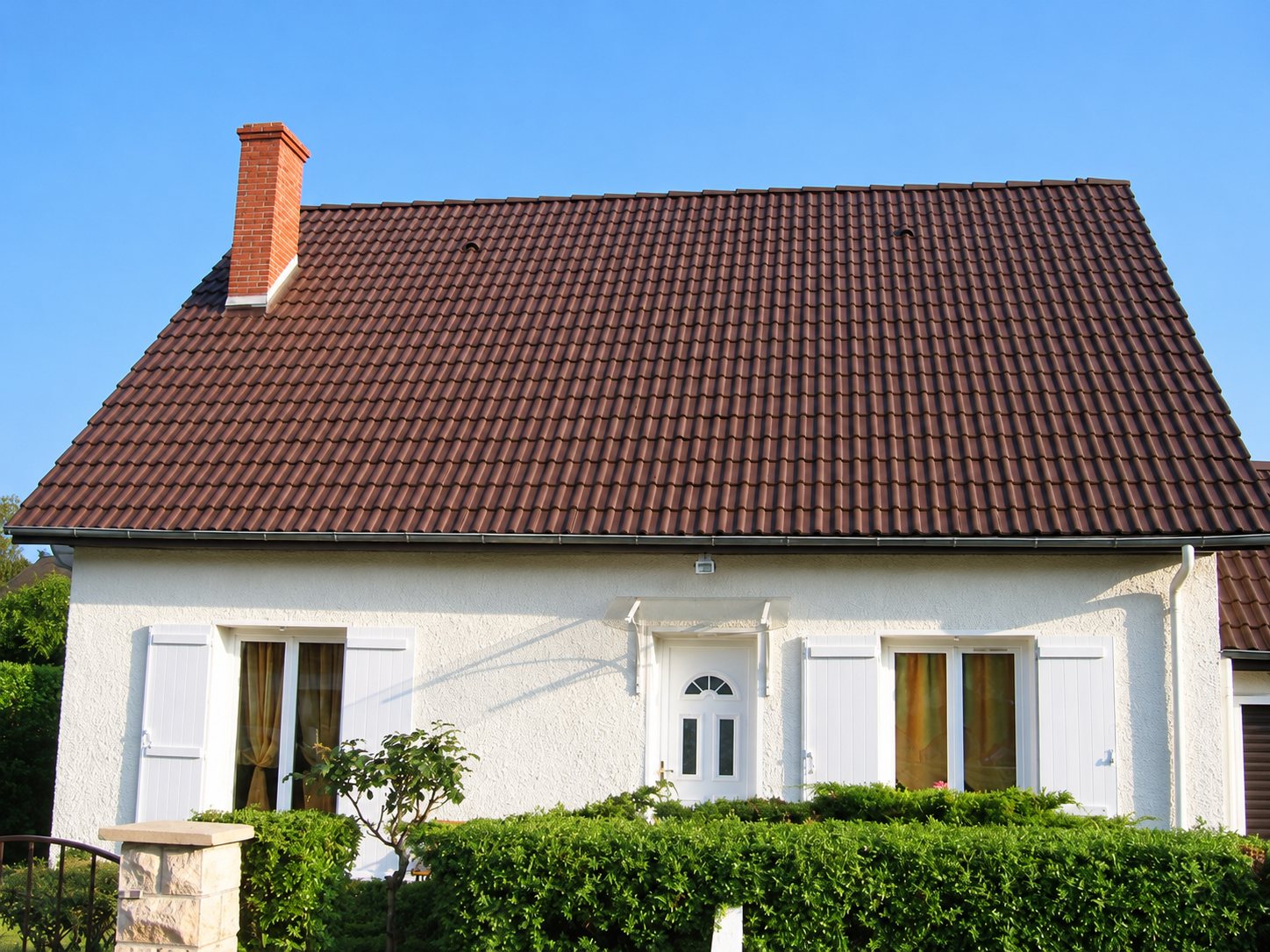 Maison individuelle à façade crépie blanche, volets blancs et toit en tuiles brunes surmonté d'une cheminée en briques, derrière une haie verte.