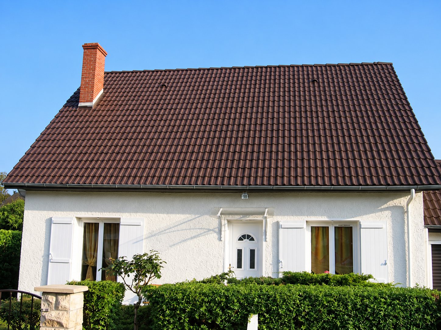 Façade d'une maison aux murs blancs crépis, surmontée d'un toit en tuiles brunes avec une cheminée en briques, derrière une haie verte.