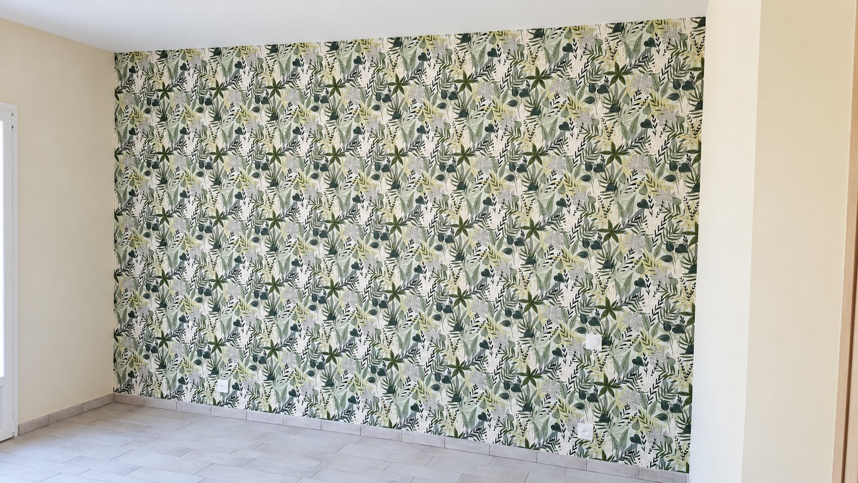 Un mur recouvert de papier peint à motifs de feuillages verts et blancs dans une pièce vide aux murs clairs et au carrelage gris.