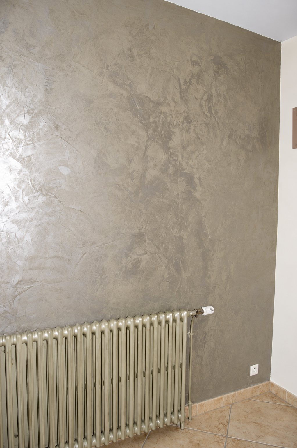 Un mur de pièce recouvert d'un enduit décoratif gris nacré aux effets métallisés, avec un radiateur en fonte peint en beige au pied du mur.