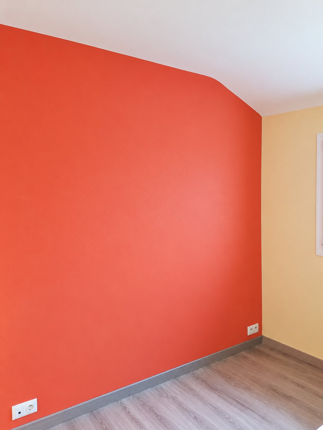 Une pièce sous combles rénovée avec un mur d'accent orange, des murs beige clair, des plinthes grises et un parquet clair.