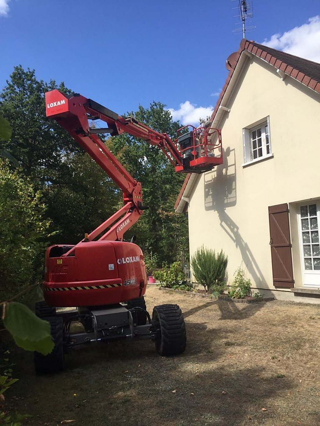 Une nacelle élévatrice articulée rouge de marque Loxam déploie son bras vers le toit d'une maison beige, avec un opérateur dans la plateforme.