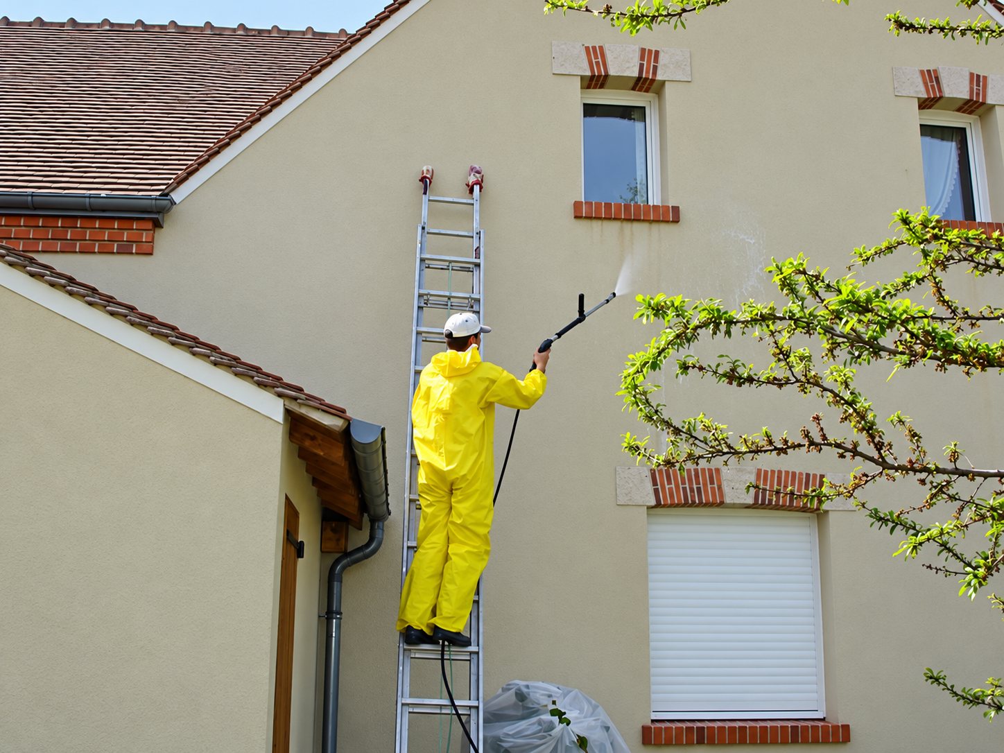 Un ouvrier en combinaison jaune, monté sur une échelle, nettoie la façade d'une maison avec une lance haute pression.