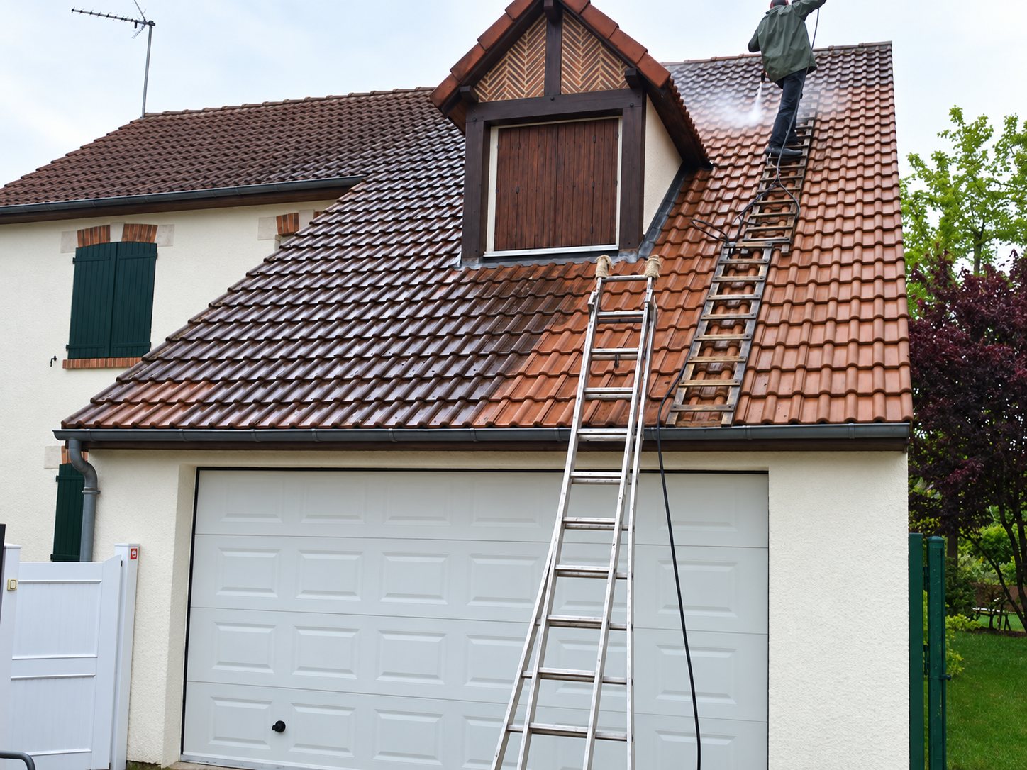 Un ouvrier nettoie au jet haute pression le toit en tuiles d'une maison, dont la moitié nettoyée apparaît plus claire, avec deux échelles posées contre le garage.