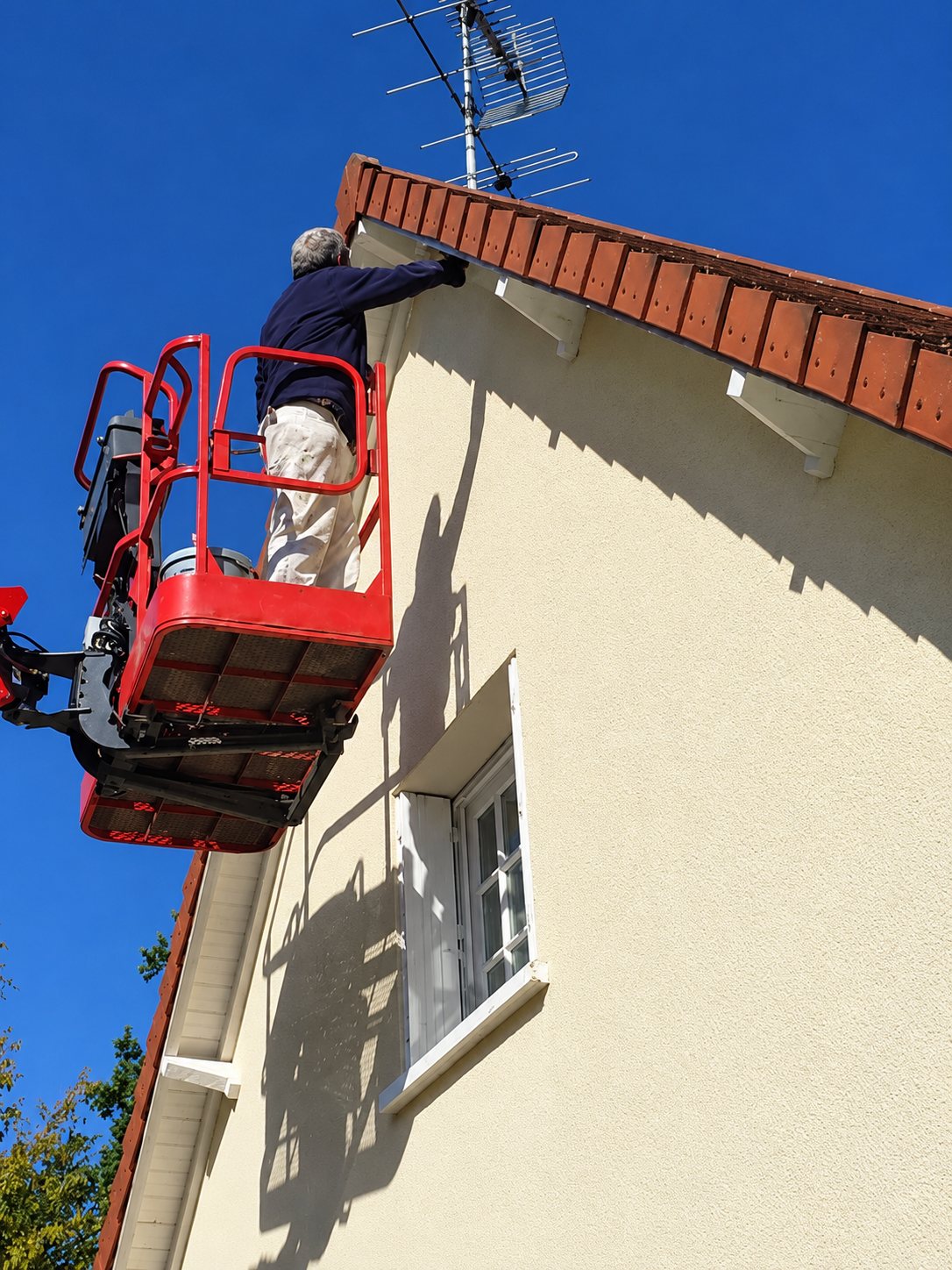 Un ouvrier debout dans une nacelle élévatrice rouge intervient sous le débord du toit d'une maison à façade crépie beige, sous un ciel bleu.