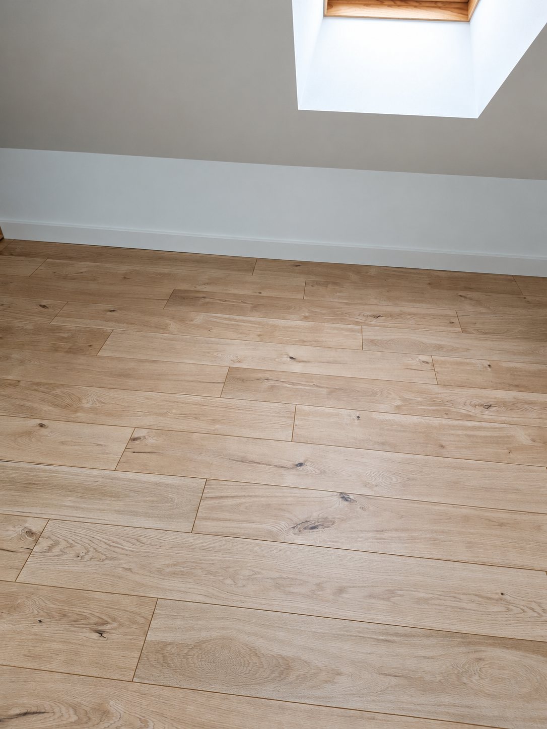 Sol en lames de parquet stratifié couleur chêne clair posé dans une pièce sous les combles, avec une fenêtre de toit lumineuse en haut de l'image.