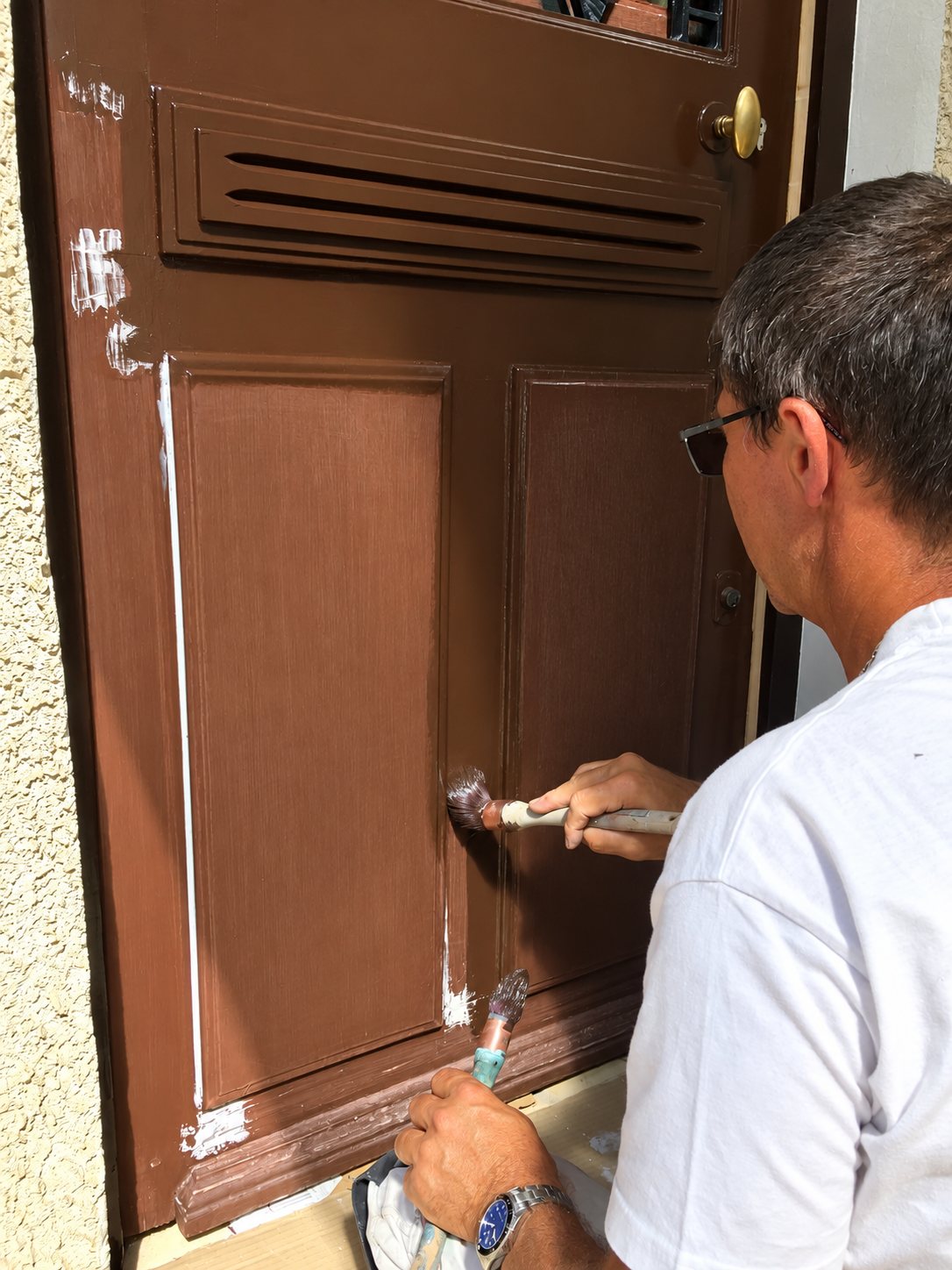 Un peintre en t-shirt blanc peint au pinceau une porte d'entrée en bois à panneaux avec de la peinture marron.