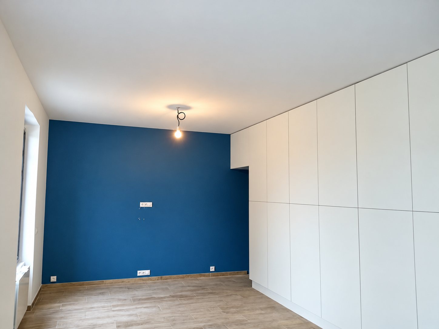 Pièce vide fraîchement rénovée avec un mur bleu foncé, de grands placards blancs sur toute la hauteur, un sol imitation bois et une ampoule suspendue.
