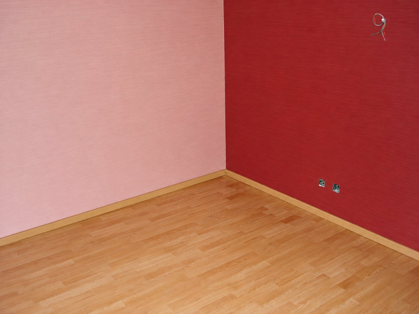 Coin d'une pièce vide avec un mur rose clair, un mur bordeaux, des plinthes en bois et un sol stratifié clair.