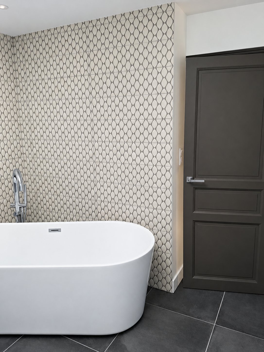 Salle de bains rénovée avec une baignoire ovale blanche, un robinet chromé sur colonne, un carrelage mural à motifs gris et blanc, une porte grise et un sol en carreaux anthracite.