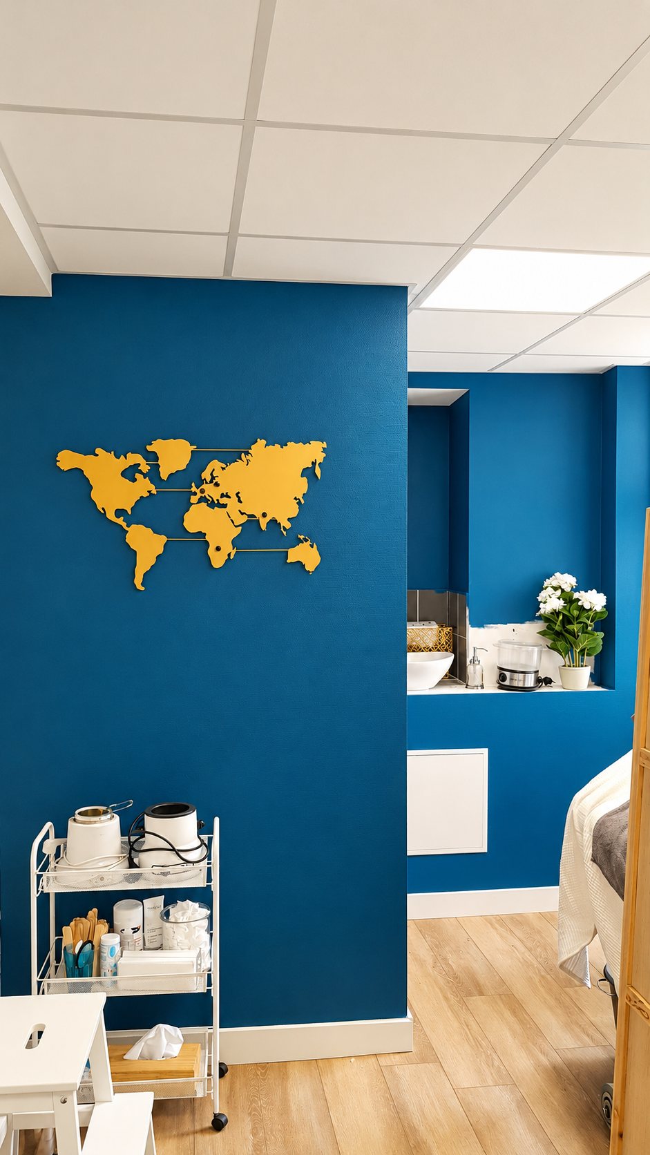 Intérieur d'une cabine de soins aux murs bleu canard, avec une carte du monde dorée au mur, une desserte blanche à roulettes, un lavabo et une table de massage.