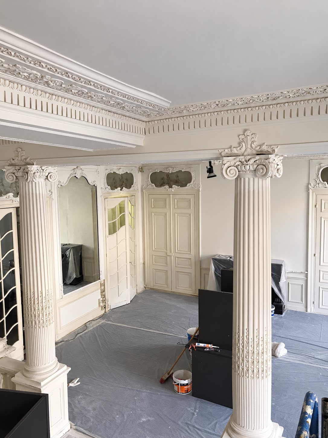 Grand salon ancien en cours de rénovation, avec colonnes cannelées, corniches en plâtre sculptées, portes blanches et sol recouvert de bâches de protection.