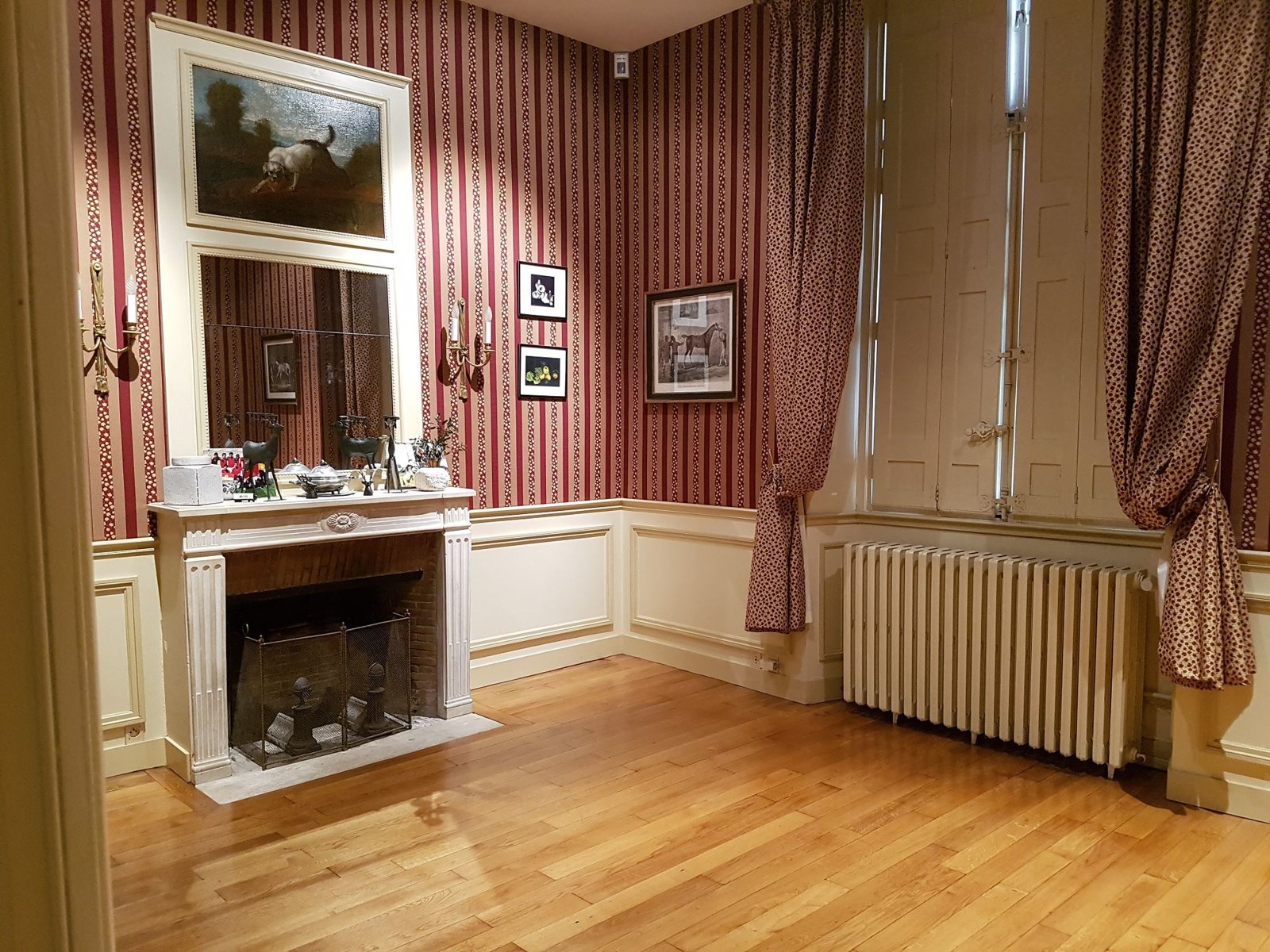 Pièce élégante aux murs rayés rouge et crème, avec cheminée surmontée d'un miroir, radiateur en fonte, rideaux à motifs et parquet en bois clair.