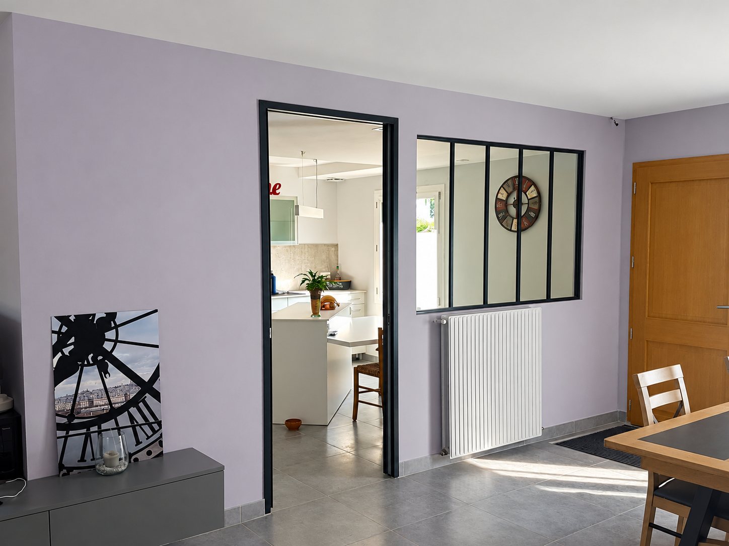 Un salon aux murs mauves dont une verrière d'atelier noire et une porte ouvrent sur une cuisine blanche, avec un radiateur vertical et un carrelage gris.