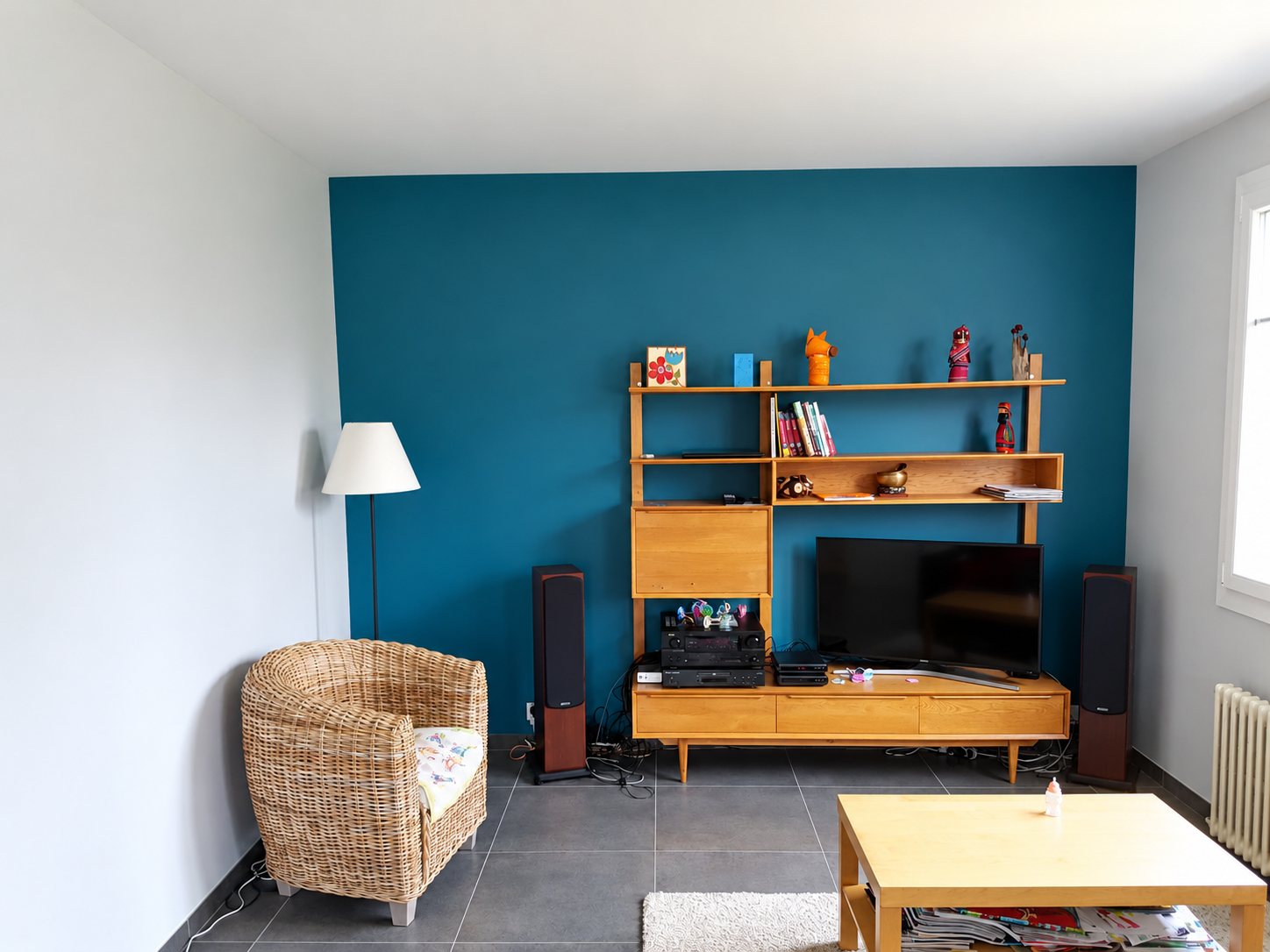 Un salon lumineux dont le mur du fond est peint en bleu canard, avec un meuble télévision en bois, un fauteuil en rotin et une table basse.