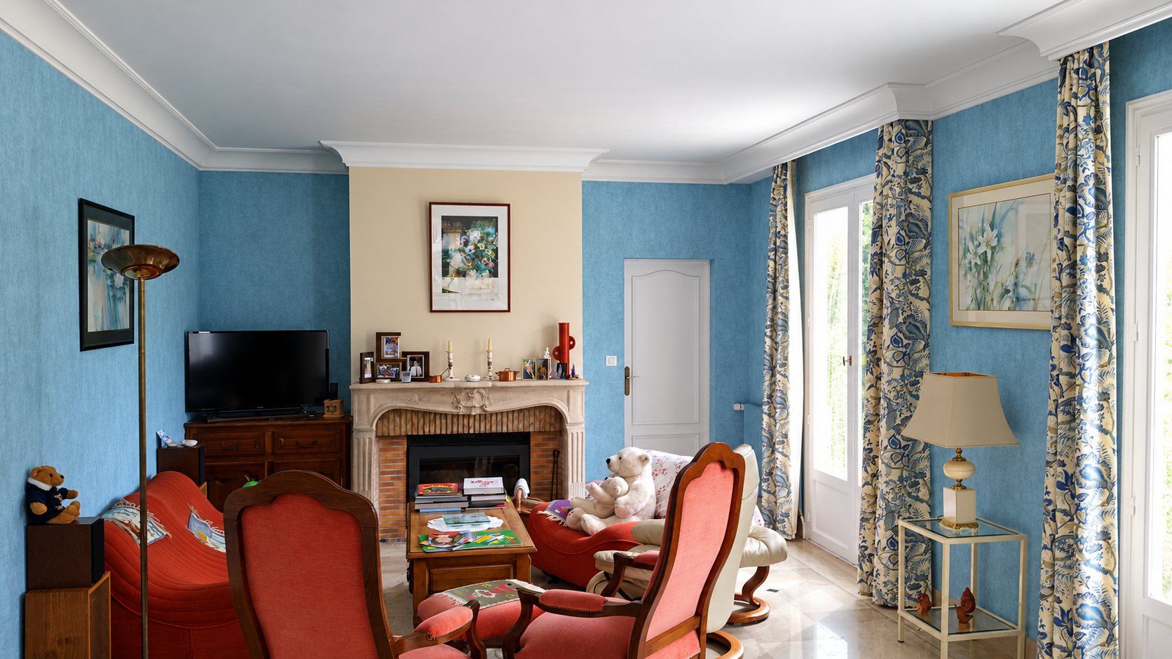 Un salon aux murs bleus texturés avec une cheminée en pierre, des fauteuils rouges, des rideaux à motifs bleus et crème et des portes-fenêtres lumineuses.