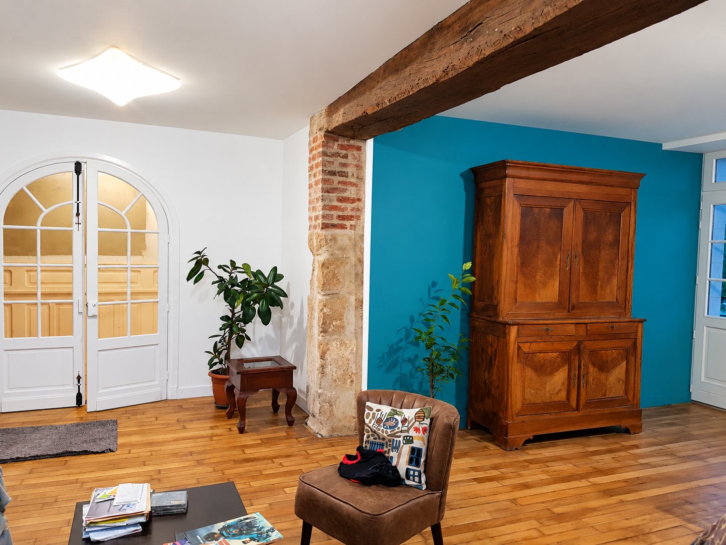 Salon rénové avec parquet, poutre en bois, pilier de briques, mur bleu canard, armoire ancienne et porte vitrée blanche cintrée.