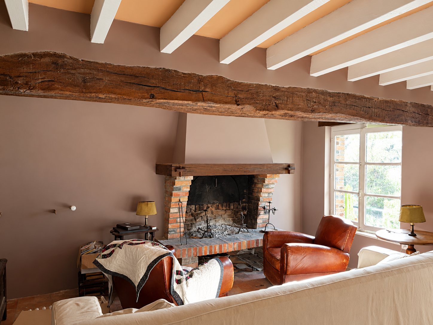 Un salon aux murs rosés avec une cheminée en briques, de grosses poutres en bois, deux fauteuils club en cuir et un canapé clair.