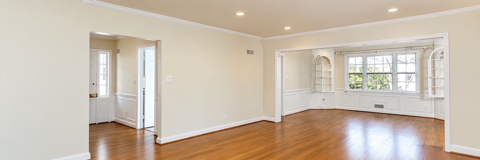 Pièce vide et lumineuse avec murs beige clair, boiseries blanches, grandes fenêtres, étagères intégrées et parquet en bois foncé.