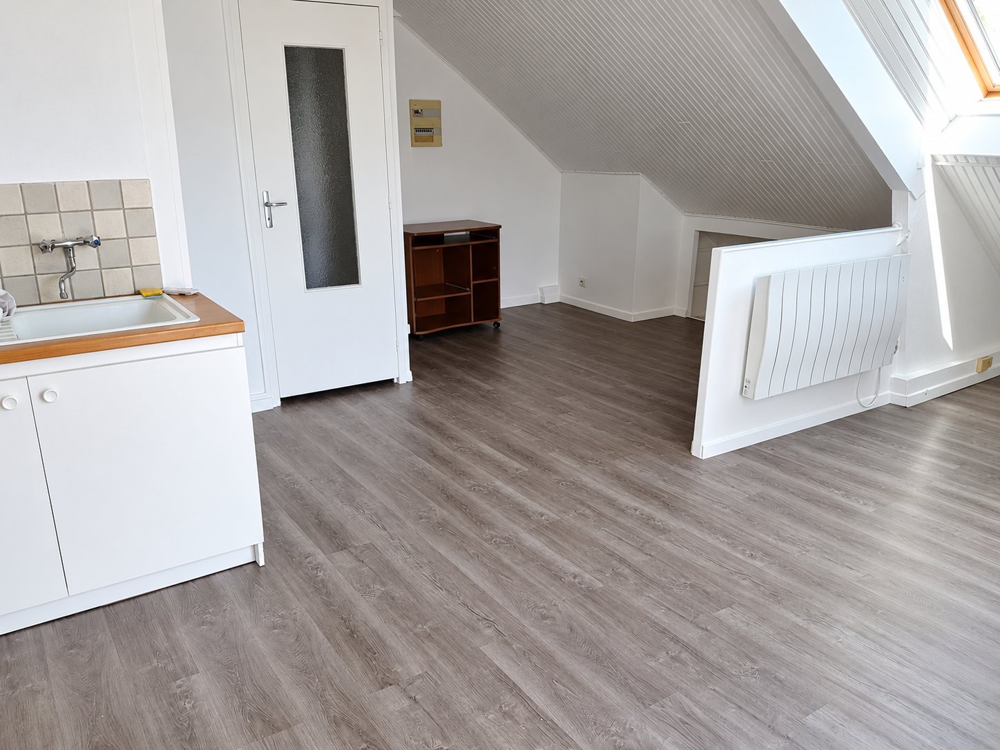 Pièce mansardée aux murs blancs avec un sol stratifié gris neuf, un coin cuisine avec évier, une porte vitrée et un radiateur électrique.